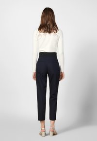 En kvinna står med ryggen mot betraktaren, klädd i en vit topp och marinblå byxor, vilket avslöjar en stilfull och detaljerad spetsdesign på hennes topp.