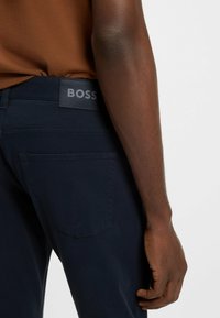 Marineblauwe broek met een gladde textuur, voorzien van een achterzak en een grijze "BOSS" logo label. Zichtbare stiksels omlijnen de zakken.
