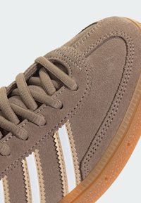 Brūni zamšādas sneakers ar baltām zick-zack svītrām, plakanu auklu un teksturētu gumijas zoli. Tuvplāns izcelt materiālu un dizainu.