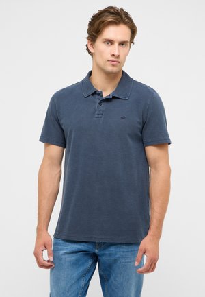 Junger Mann, der ein dunkelblaues kurzärmeliges Poloshirt und blaue Jeans trägt, vor einem schlichten grauen Hintergrund stehend.