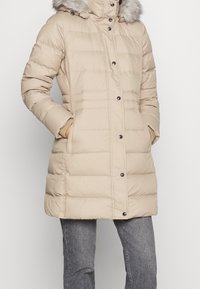 Beige gewatteerde jas met een met bont gevoerde capuchon, drukknoopsluiting, zijzakken en ribgebreide manchetten. Lichtgewicht stof met een doorgestikt patroon.