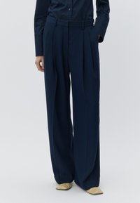 Calças azul-marinho de perna larga com pregas, feitas de tecido liso. Combinadas com uma camisa escura e sapatos bege, apresentam uma cintura ajustada.