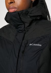 Svart jacka med hög krage och dragkedja fram, har en strukturerad design, Columbia-logotyp och justerbara snoddar.