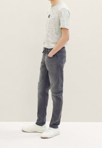 Persona in piedi con le mani in tasca, che indossa una camicia a maniche corte grigia chiara, jeans grigio scuro e sneakers bianche su uno sfondo uniforme.