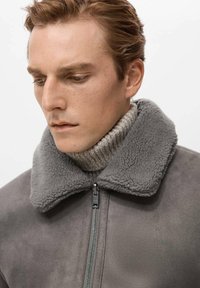 Grijze shearlingjas met een grote kraag, ritssluiting en een getextureerde wollen coltrui eronder, met een gladde stoffen afwerking.