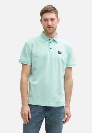 Polo shirt vert menthe avec un col, patte de boutonnage à trois boutons et étiquette marine. Porté avec un jean bleu. Texture lisse, manches courtes.