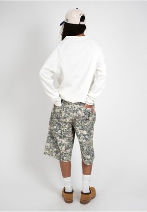 Persona de espaldas que lleva una sudadera blanca, pantalones cortos largos con estampado de camuflaje, botas marrones, calcetines blancos y una gorra blanca y azul marino.