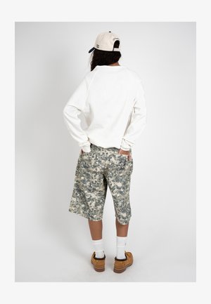 Persona de espaldas que lleva una sudadera blanca, pantalones cortos largos con estampado de camuflaje, botas marrones, calcetines blancos y una gorra blanca y azul marino.