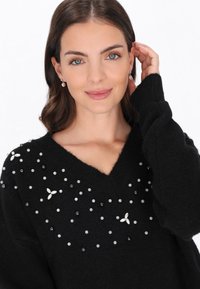 Czarny wełniany sweter z dekoltem w serek, ozdobiony różnymi koralikami i motywami kwiatowymi. Modelka dotyka swoich włosów, nosząc perłowe kolczyki w kształcie kropli.
