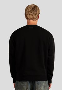 Felpa nera con colletto a girocollo e polsini a costine, dotata di un'etichetta texturizzata sul retro. Tessuto liscio e vestibilità rilassata.