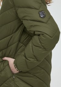 Oliven grøn pufferjakke med quiltet design, der har et sort logo-patch på skulderen og en hånd stukket ind i en sidelomme.