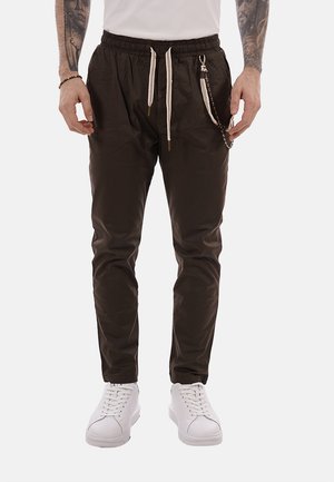 Uomo che indossa pantaloni jogger neri con coulisse bianche e un portachiavi, abbinati a scarpe da ginnastica bianche e braccia tatuate.