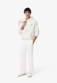Hombre vestido con sudadera con capucha blanca y pantalones, sosteniendo una prenda color beige claro sobre el hombro, de pie frente a un fondo blanco.