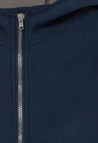 Marineblå fleecejakke med oppstående krage, hel glidelås, og merkevarenavnet "VINGINO" brodert i matchende farge på forsiden.