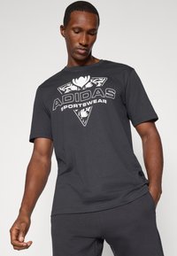 Juodasis medvilnės marškinėliai su dideliu baltu Adidas logotipu ir gėlių raštu trikampio forma priekyje. Trumpomis rankovėmis, apvali iškirptė.