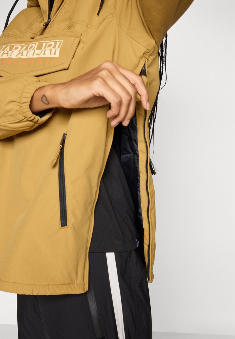 Person, der den seitlichen Reißverschluss einer senfgelben Napapijri-Jacke aufzieht, trägt schwarze Hosen mit einem weißen Streifen und ein schwarzes Shirt darunter.