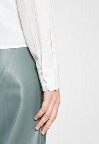 Blouse blanche à manches longues avec des poignets en dentelle et un détail de bouton, associée à un pantalon en simili cuir vert doux. Texture lisse visible.