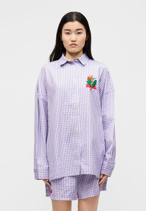 Młoda kobieta ubrana w oversize lawendową koszulę w kratę zapinaną na guziki oraz pasujące szorty z kolorową kwiatową haftem na piersi i nodze.