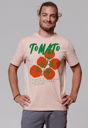 Zachte roze katoenen t-shirt met een groen en rood tomaatgrafiek en tekstontwerp op de voorkant. Korte mouwen en een ronde hals.
