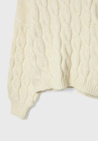 Manche et bas du corps d'un pull en maille torsadée couleur crème avec poignets et ourlet côtelés sur un fond uni.