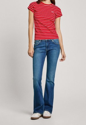 Femme portant un haut à manches courtes rouge et blanc rayé, un jean évasé bleu et des baskets blanches, debout sur un fond uni.