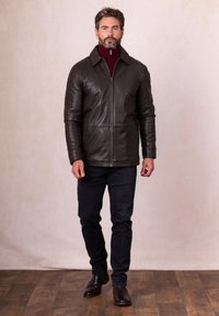 Brown leather jacket with a zip front, high collar, and two side pockets, paired with dark jeans and brown leather boots on a light background.