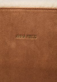 Matériau en daim marron avec une texture douce ; présente un logo doré en relief "ANNA FIELD". Le design est simple et élégant.