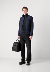 Zweigotni vodoodporna jakna s visokim ovratnikom, črna turtleneck majica, črn jeans, in črna usnjena torbica, nosena z črnimi slip-on čevlji.