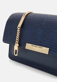 Granatowa skórzana kopertówka z fakturowaną powierzchnią, złotym łańcuszkowym paskiem oraz metalową tabliczką z wygrawerowaną nazwą „Michael Kors”.