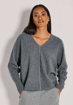 Vrouw met lang stijl haar, gekleed in een grijze V-hals trui met een witte verticale naad aan de voorkant en bijpassende grijze broek, met de handen in de zakken.