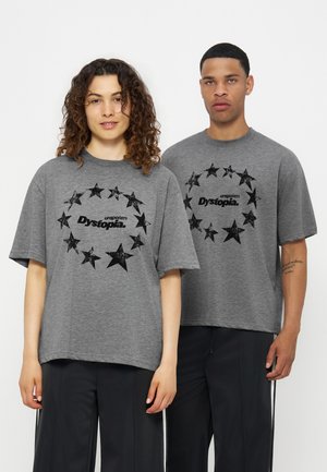 Deux personnes portent des t-shirts gris oversize avec un cercle d'étoile noir et le texte « unspoken Dystopia », associés à un pantalon noir, debout devant un fond blanc.