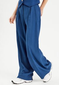 Pantalon bleu à jambes larges avec plis, fabriqué en tissu lisse. Associé à des baskets blanches, mettant en avant une coupe décontractée et un design drapé.