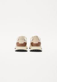 Tenisi cu o parte superioară din plasă beige, accente din suede maro și o talpă din cauciuc negru. Prezintă logo pe toc și un design cu amortizare.