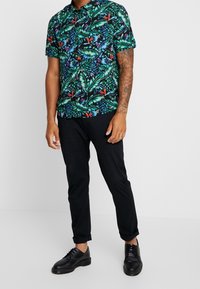 Mann in einem bunten kurzärmligen Hemd mit tropischem Muster, schwarzen hochgekrempelten Hosen und schwarzen eleganten Schuhen, der vor einem schlichten Hintergrund steht.