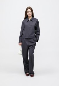 Grå, kort skjorte med spiss krage og lange ermer, kombinert med matchende vide bukser med vid legs. Modellen holder en liten hvit veske.