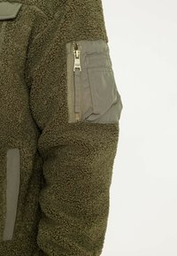 Puszysta oliwkowo-zielona polarowa kurtka z teksturowaną powierzchnią i kieszenią na zamek na lewym ramieniu, z gładkim, kontrastowym akcentem z materiału.