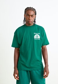 Grøn bomulds t-shirt med en løs pasform, der har en rund halsudskæring og grafik på brystet. Kombineret med matchende grønne shorts.