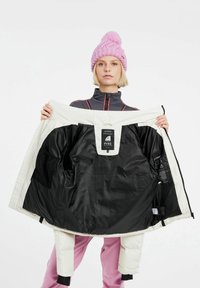 Witte geïsoleerde jas met zwarte voering; heeft een label van binnen, een ritssluiting en elastische manchetten. Het model houdt de jas open, waardoor het interieur zichtbaar is.