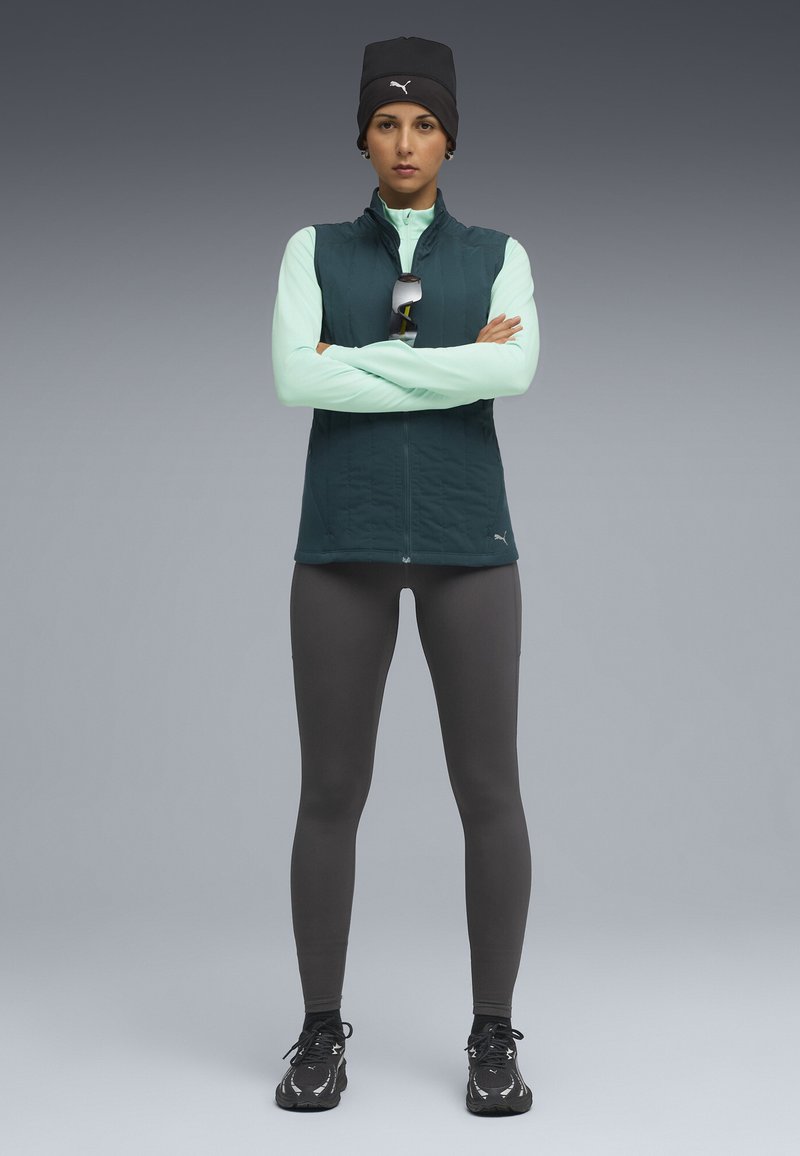 Chaleco acolchado color teal sobre una camiseta de manga larga verde claro, combinado con leggings oscuros y zapatillas deportivas negras. Un gorro negro y gafas de sol completan el atuendo.