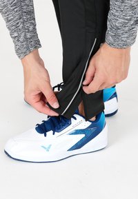 Vita och blåa sportskor med texturerade detaljer. Svarta byxor med blixtlås vid vristerna och en grå långärmad tröja, händer som justerar byxbenens nederkant.