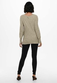 Beżowy sweter dzianinowy z długimi rękawami i plecami w kształcie V, zestawiony z czarnymi slim fit jeansami i czarnymi szpilkami. Gładka struktura.