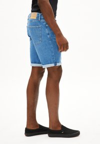 Shorts en denim bleu clair avec un ourlet retroussé, dotés de poches arrière et d'une étiquette en cuir beige sur la taille. Chaussures noires à enfiler visibles.