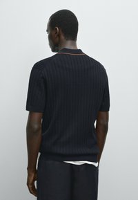 Maglione corto a maniche corte a coste navy con scollatura a girocollo e dettaglio marrone al colletto, indossato sopra una camicia bianca, mostrato dal retro.