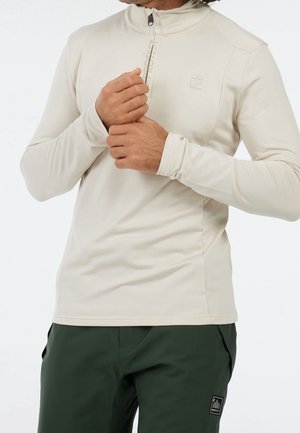 Mężczyzna w beżowej koszulce z długim rękawem i suwakiem do połowy oraz ciemnozielonych spodniach z małą naszywką z logo na nodze, poprawia rękaw.