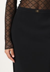 Saia preta lápis em um tecido suave, apresentando um detalhe de logotipo dourado. A blusa de manga longa tem uma malha preta translúcida com padrão de diamante.