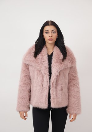 Kvinde med langt mørkt hår iført en lyserød, fluffy faux pelsjakke over sort tøj, stående mod en ensfarvet lys baggrund.