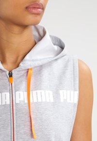 Šedá bezrukávová mikina s kapucí s nastavitelným šňůrkovým stahováním, s bílým nápisem "PUMA" a oranžovými akcenty na zipu pro lepší viditelnost.