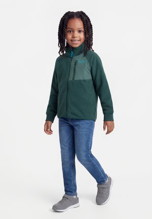 Usmievajúce sa dieťa s vrkočmi, oblečené v tmavozelenej bunde na zips, modrých džínsach a sivých teniskách, stojace pred bielym pozadím.