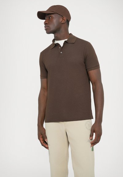 Hnedá polokošeľa s krátkym rukávom s textúrovaným úpletovým vzorom, zapínaním na tri gombíky a jednoliatym golierom, v kombinácii s svetlobéžovými nohavicami.