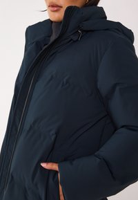 Doudoune bleu marine avec capuche, dotée d'une fermeture éclair frontale, de poches latérales et d'une texture matelassée pour la chaleur et le confort.